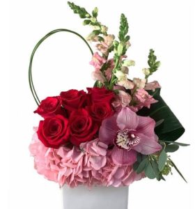 An arrangement of pink hydrangeas, red roses, pink cymbidium orchids, pink snapdragons, seeded eucalyptus, green aspidistra leaves and bear grass. 